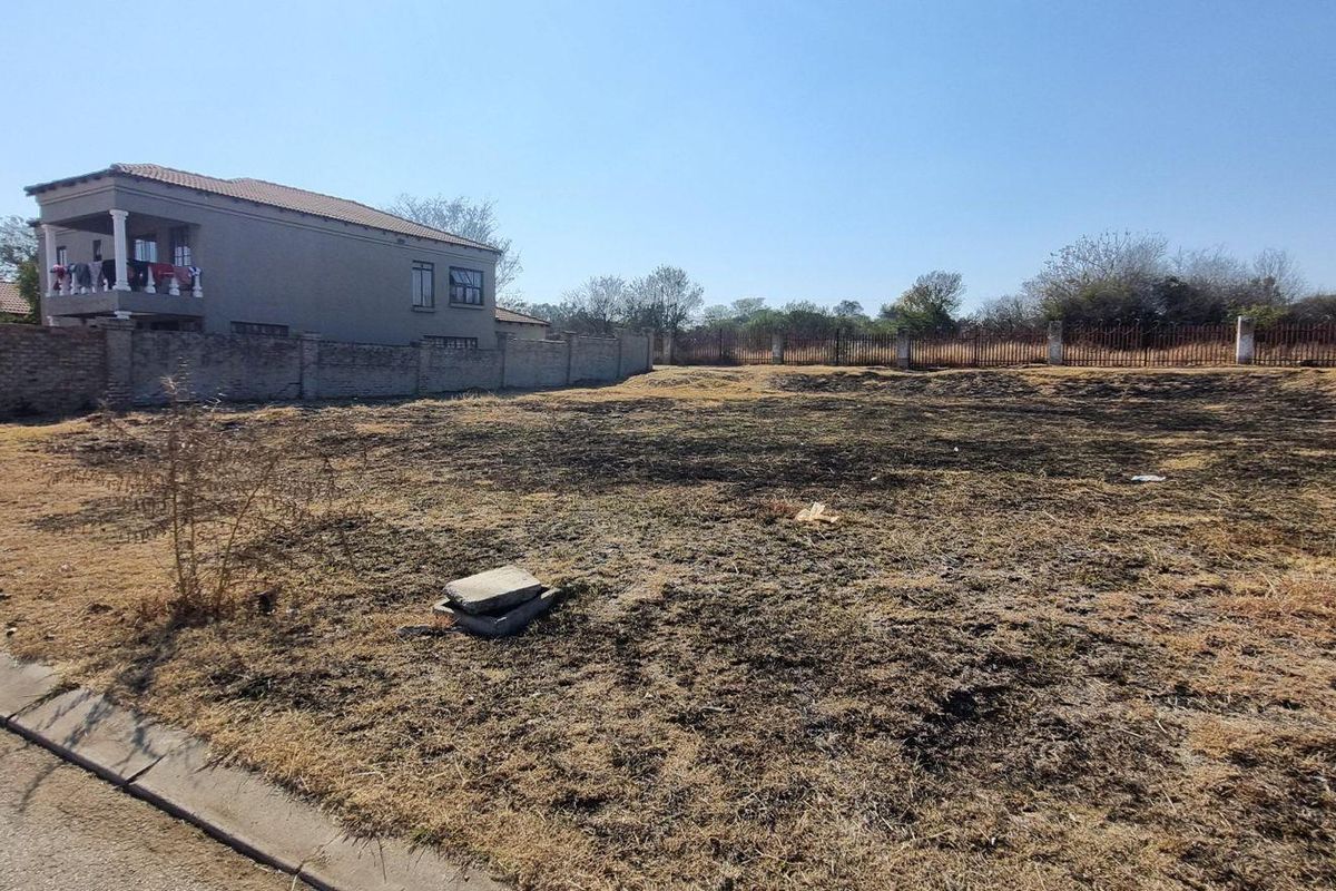 A vacant residential plot featuring a perimeter brick wall and metal fencing, situated along a paved street in a developed neighborhood.