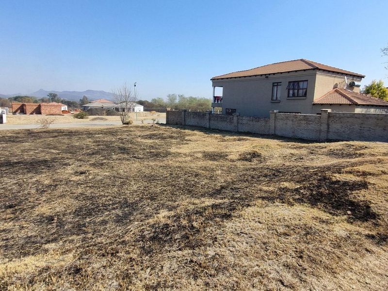 A vacant residential plot featuring dry, burnt grass with an existing perimeter wall and surrounding neighborhood houses in view.
