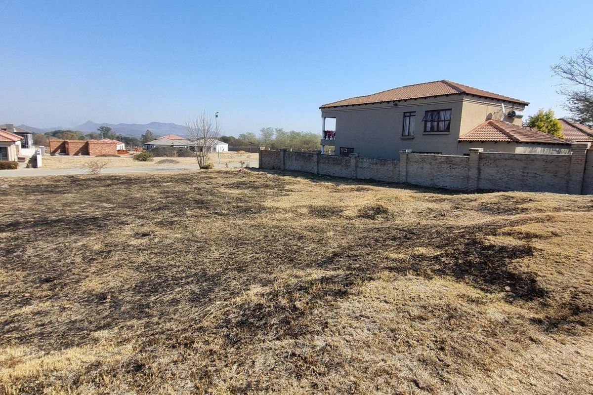 A vacant residential plot featuring dry, burnt grass with an existing perimeter wall and surrounding neighborhood houses in view.