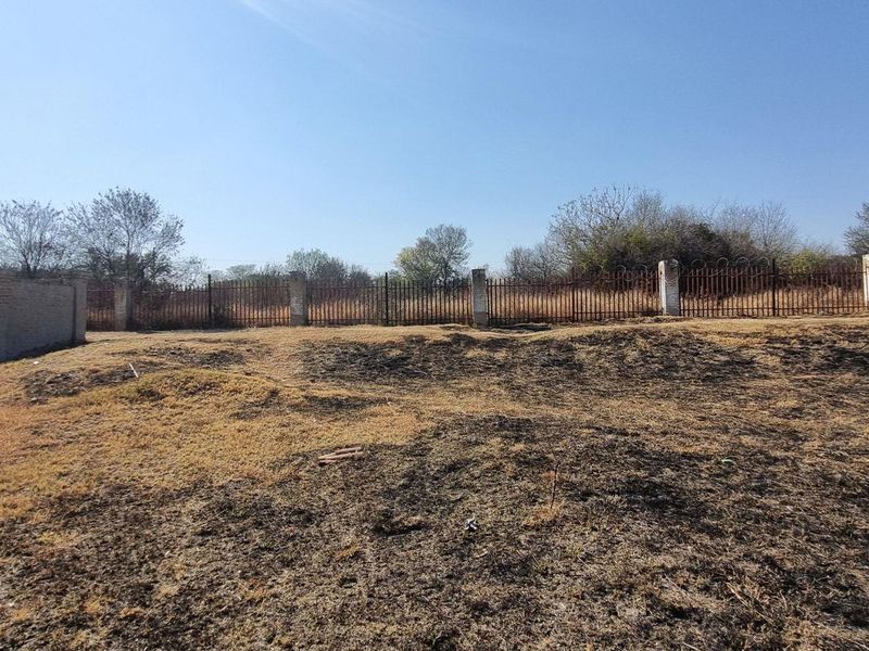 A vacant residential plot featuring perimeter brick walls and metal security fencing, with dry, unmaintained ground cover.