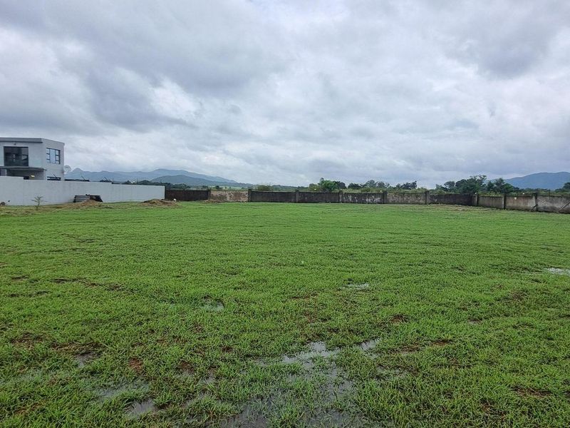 A spacious, level residential plot featuring a grass lawn and perimeter boundary wall, situated in a developing neighborhood with mountain views.