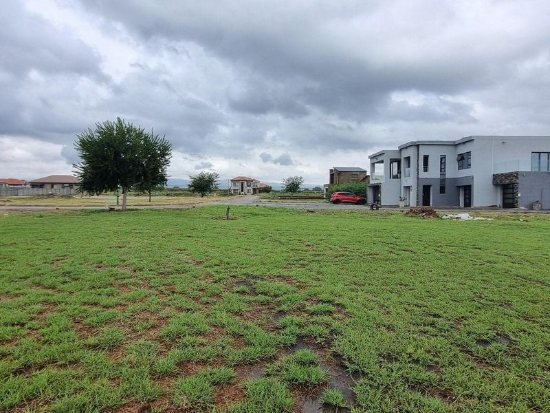 A vacant residential plot featuring a grass lawn with surrounding neighborhood development and street access.