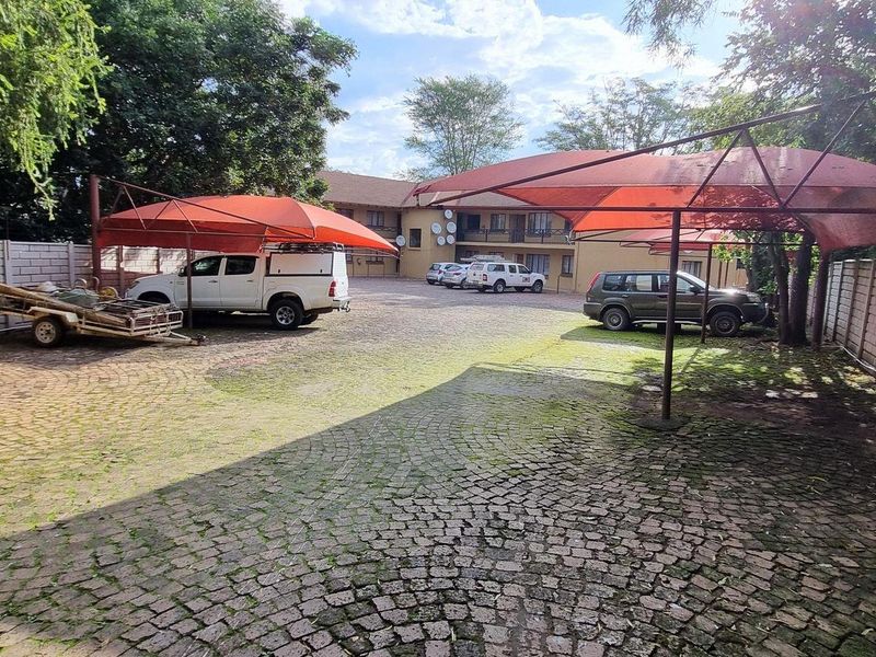 Exterior view of the apartment complex parking area featuring cobblestone paving and multiple shade cloth carports enclosed by a precast concrete boundary wall.