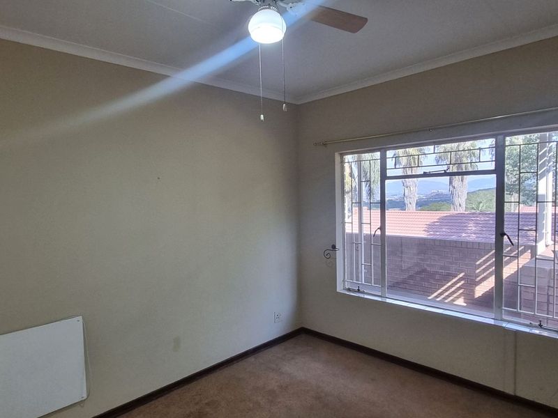 An empty bedroom featuring a ceiling fan, wall-mounted heater, and large window with security bars, offering a view of the surrounding area.