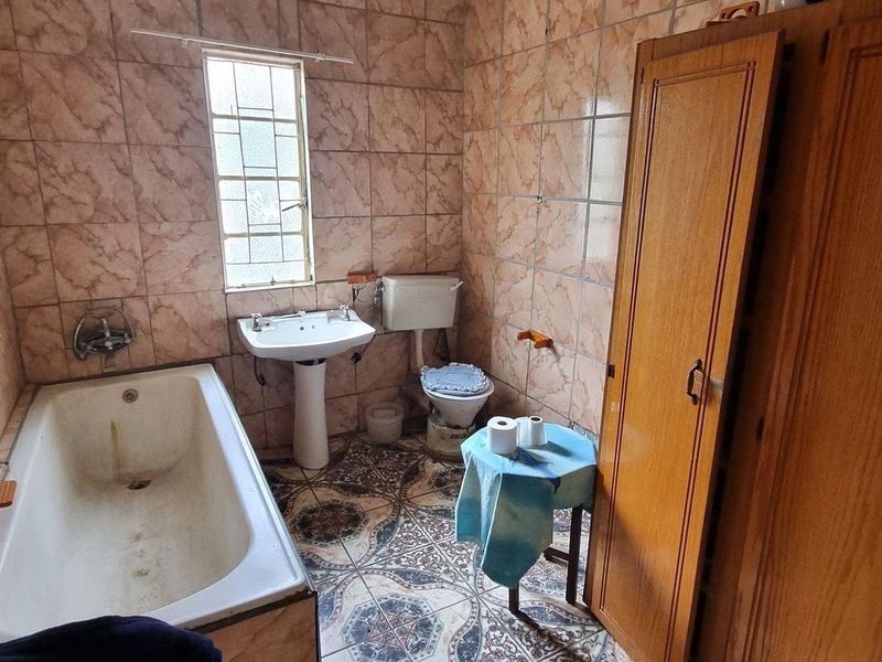 A dated bathroom featuring a bathtub, pedestal basin, toilet, and built-in wooden storage, finished with patterned floor tiles and full-height wall tiling.