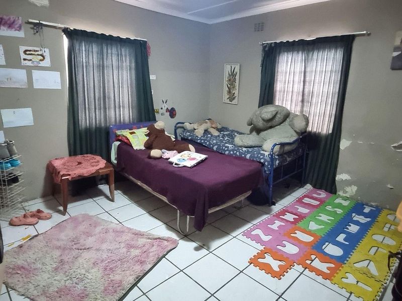A bedroom featuring tiled flooring and two windows with curtain rails. The walls show significant signs of peeling paint and plaster damage.