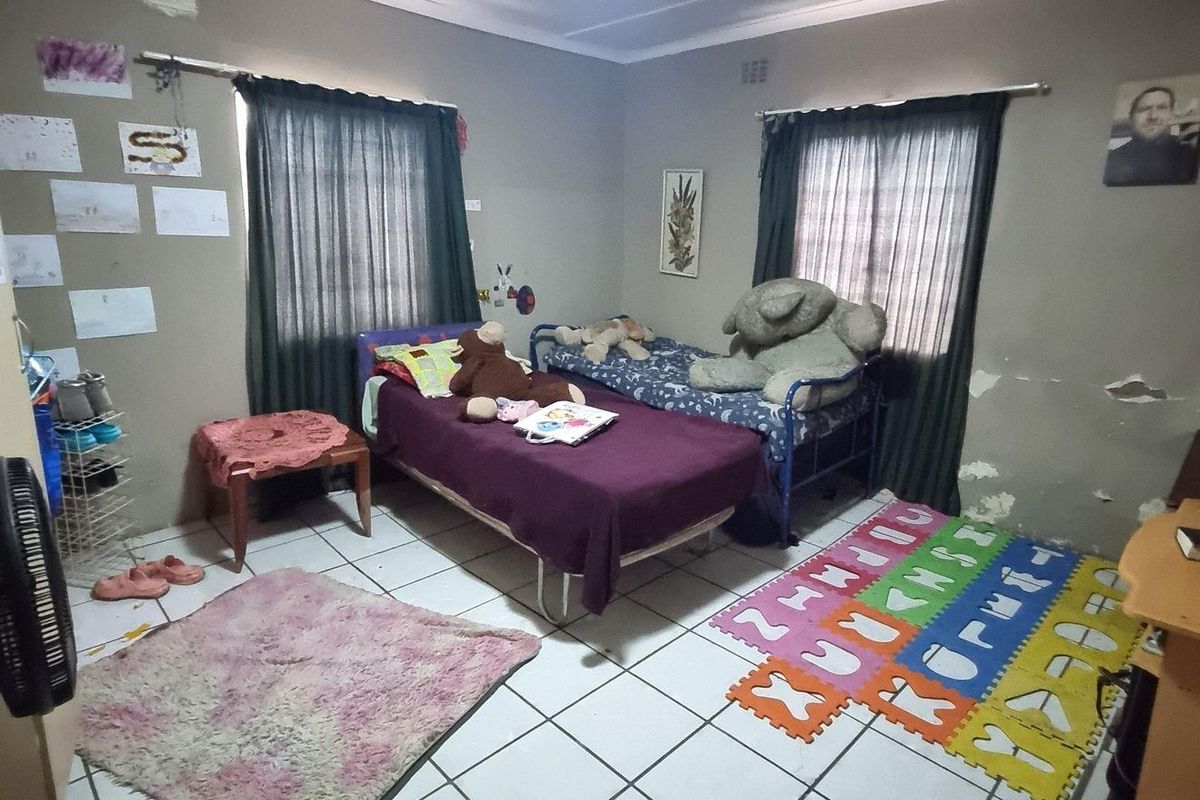 A bedroom featuring tiled flooring and two windows with curtain rails. The walls show significant signs of peeling paint and plaster damage.