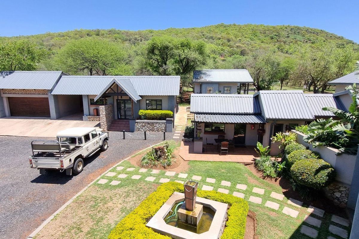 An elevated view of a farm property featuring multiple buildings with metal roofs, stone accents, a paved driveway, and a landscaped garden with a fountain.