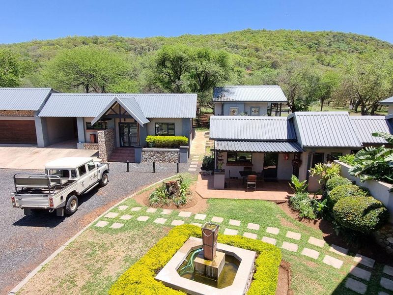 An elevated view of a farm property featuring multiple buildings with metal roofs, stone accents, a paved driveway, and a landscaped garden with a fountain.