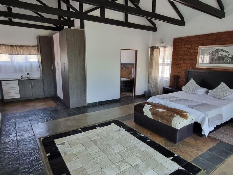 Spacious bedroom featuring exposed timber roof trusses, a rustic brick feature wall, slate flooring, and integrated built-in cupboards with en-suite bathroom access.