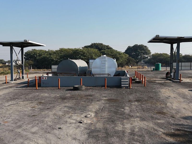 Commercial fuel depot area featuring two large storage tanks within a containment bund, protected by safety bollards and covered by dual pump canopies.