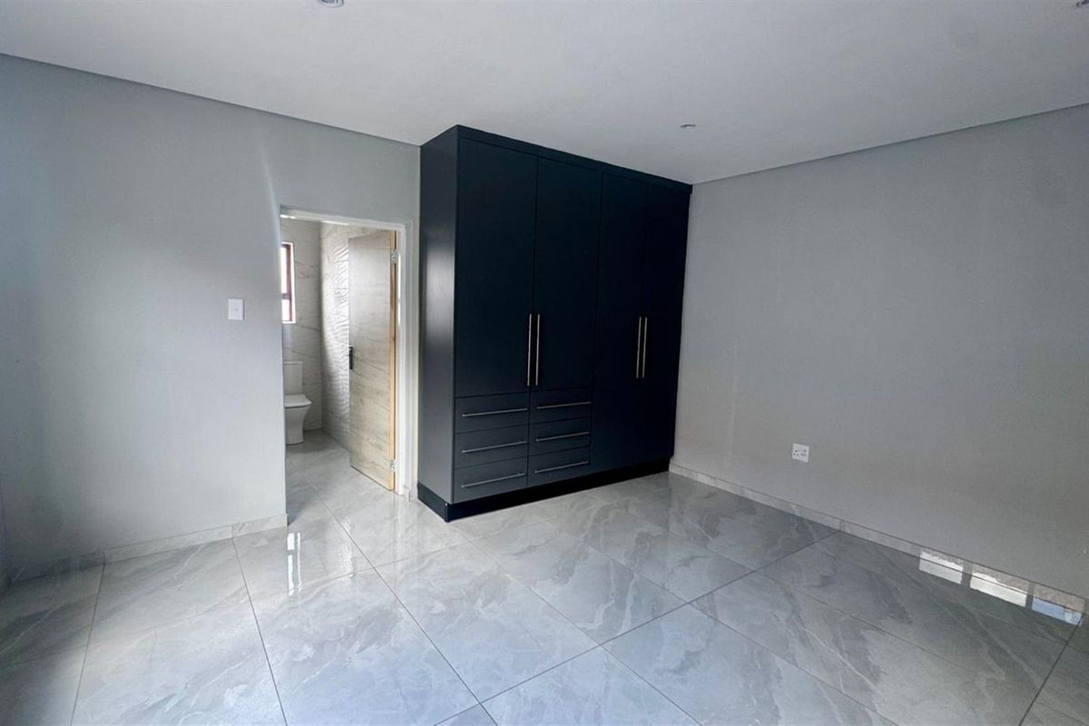 A modern bedroom featuring large-format porcelain floor tiles, neutral grey walls, and a built-in dark cabinetry unit, with an adjoining en-suite bathroom.