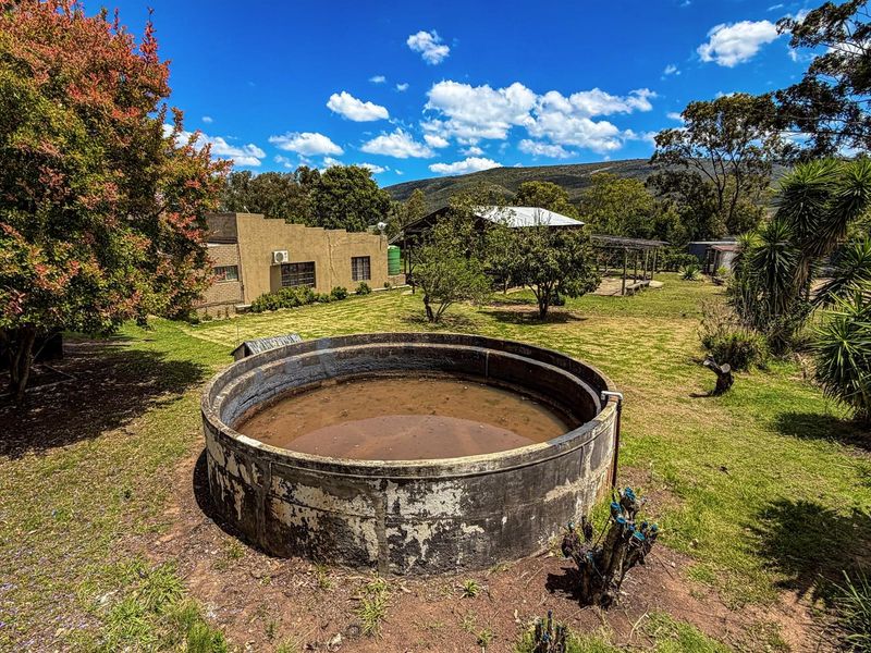 A rural farm property featuring a large concrete water reservoir in the foreground, set against a backdrop of outbuildings and mountain views.