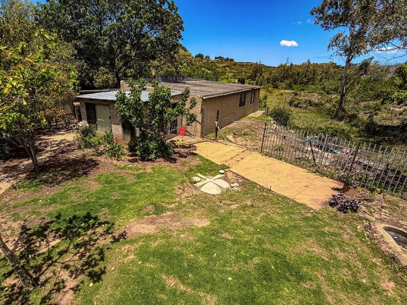 An elevated view of a farm property featuring a modest dwelling with a flat roof, surrounded by a garden and rural landscape.
