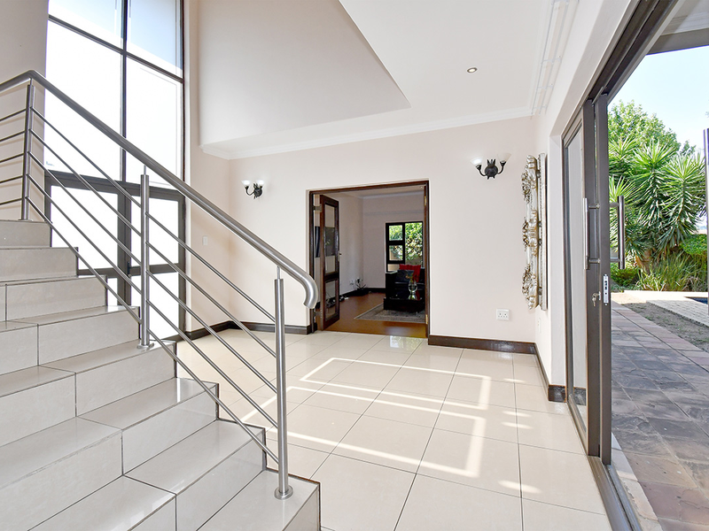 Spacious double-volume entrance hall featuring a tiled staircase with stainless steel balustrades, large windows, and direct access to the outdoor patio area.