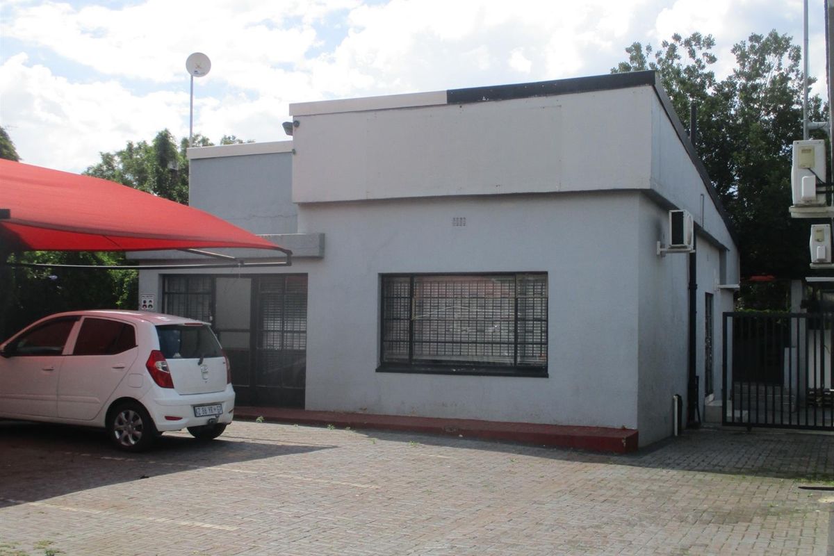 Front exterior view of a commercial property featuring a paved parking area, shade cloth carport, and security features including window bars and a metal gate.