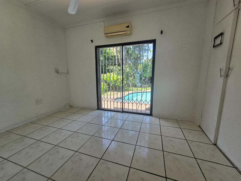 An empty living room featuring tiled floors, a wall-mounted air conditioner, and a sliding glass door with a security gate leading to the pool area.