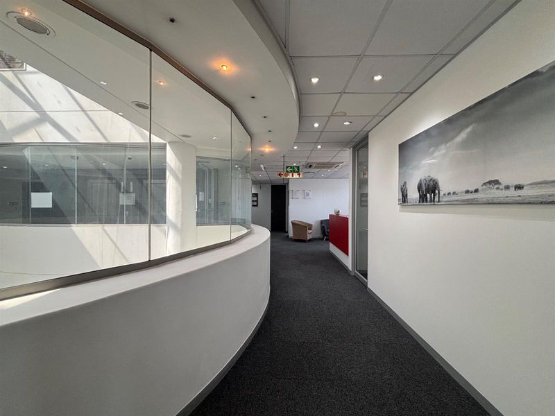 A modern office hallway featuring a curved glass partition, recessed lighting, and professional carpet flooring, providing a clean and well-maintained circulation space.
