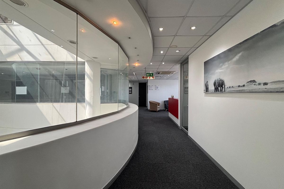 A modern office hallway featuring a curved glass partition, recessed lighting, and professional carpet flooring, providing a clean and well-maintained circulation space.