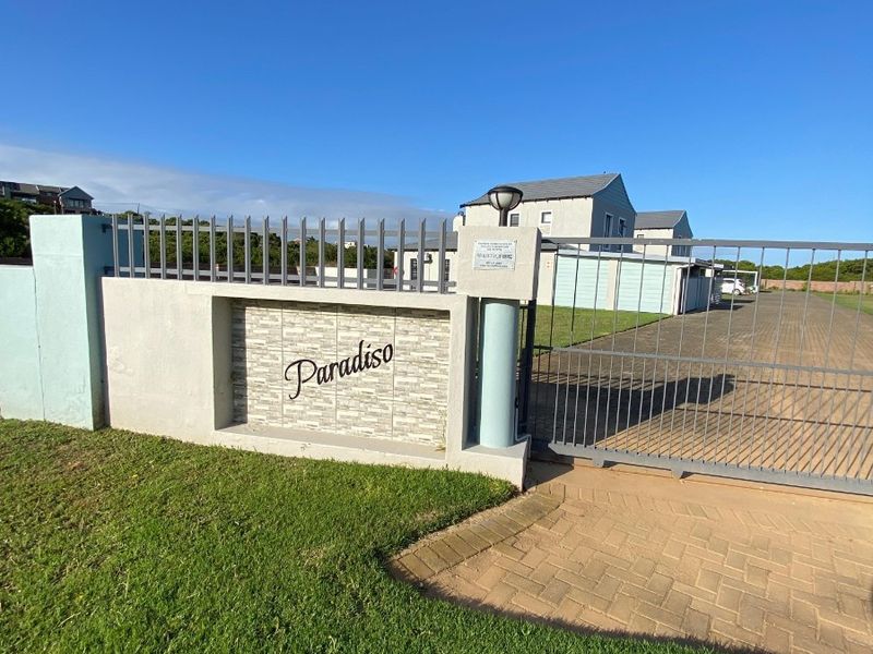 Front entrance view of the Paradiso property featuring a secure metal gate, masonry boundary wall with stone cladding, and a paved driveway.