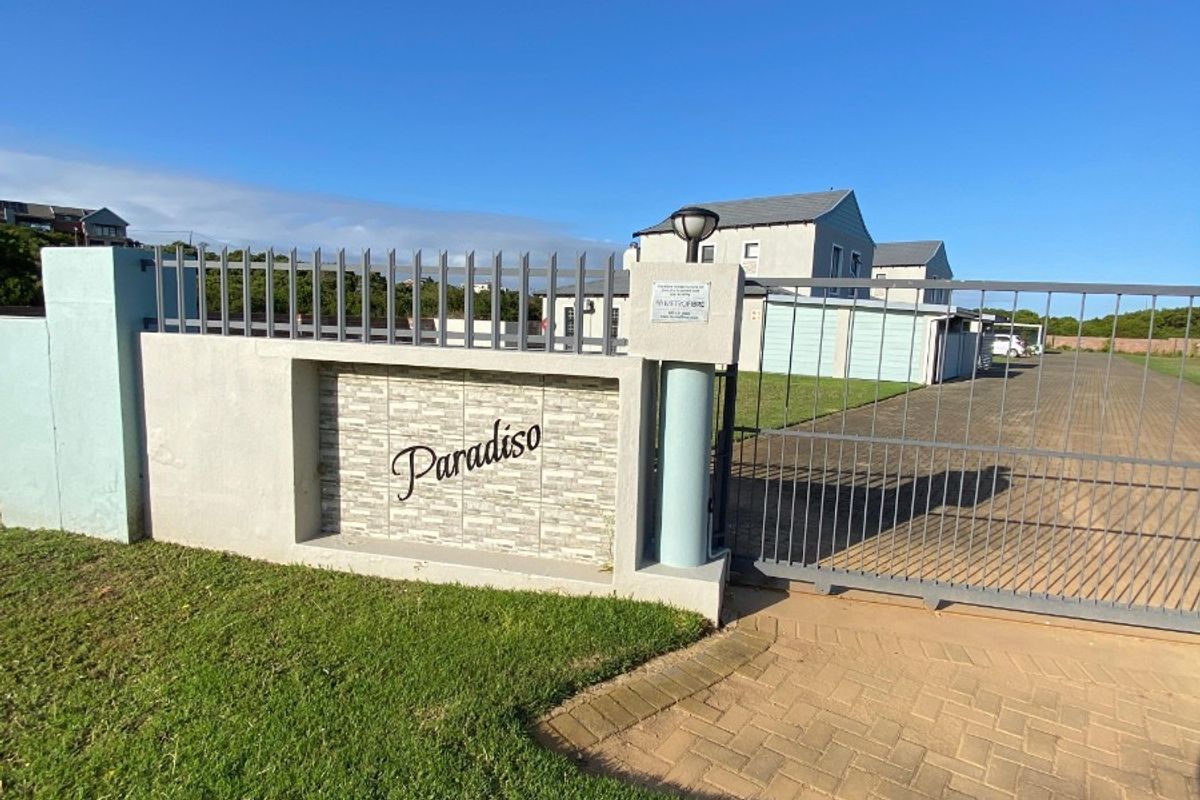 Front entrance view of the Paradiso property featuring a secure metal gate, masonry boundary wall with stone cladding, and a paved driveway.