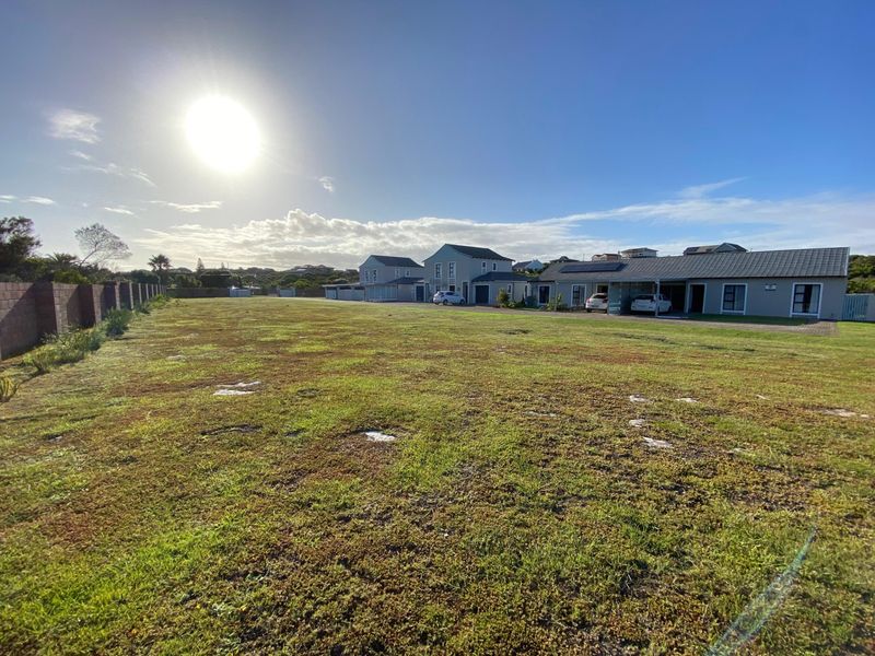 A wide-angle view of a vacant residential plot featuring a grassy landscape, boundary wall, and adjacent residential properties with carports.