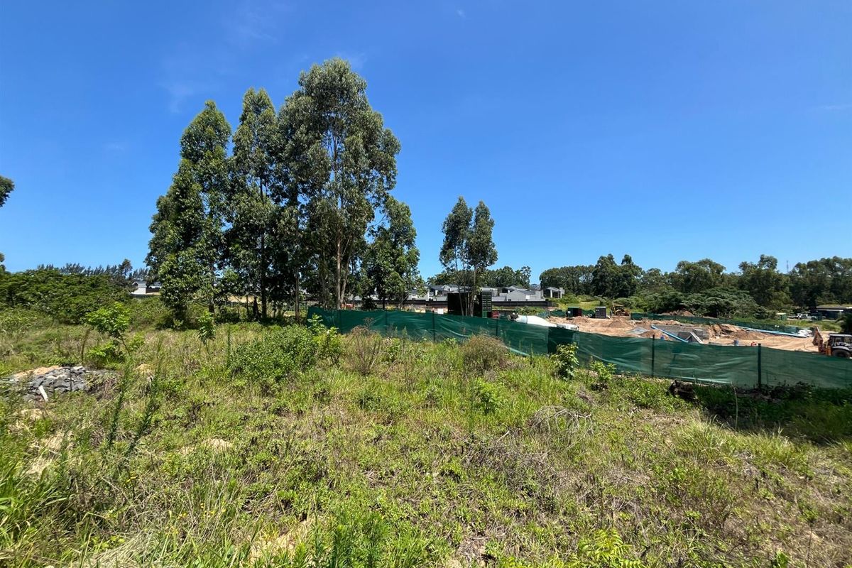 A view of a vacant residential plot featuring natural vegetation, mature trees, and temporary construction fencing surrounding a cleared area.