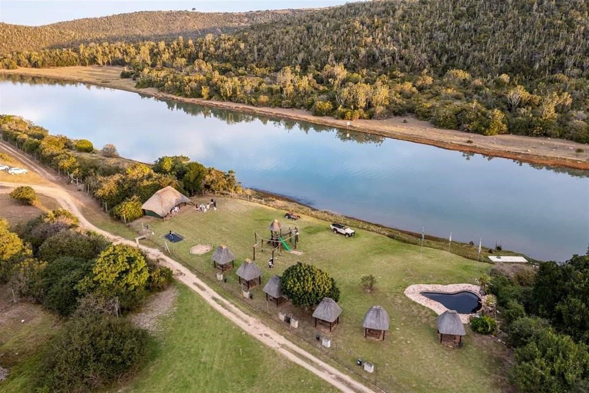 Aerial view of a riverside farm property featuring multiple thatched-roof structures, a small pool, and expansive landscaped grounds along the water's edge.