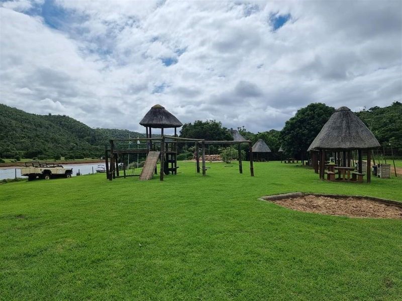 Expansive outdoor recreational area featuring thatched gazebos, a wooden playground structure, and a sandpit, set against a scenic landscape with water frontage.