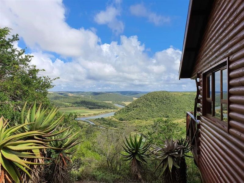 A scenic view overlooking a lush river valley and rolling hills, captured from the wooden deck of a rustic log cabin.