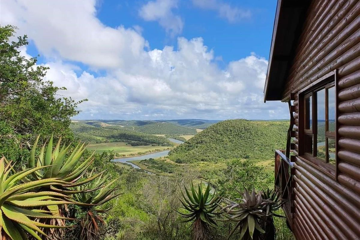 A scenic view overlooking a lush river valley and rolling hills, captured from the wooden deck of a rustic log cabin.