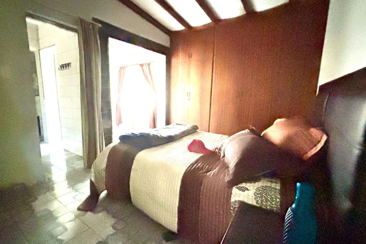 A bedroom featuring wood-paneled walls, exposed ceiling beams, and tiled flooring, with an adjacent doorway leading to another room.