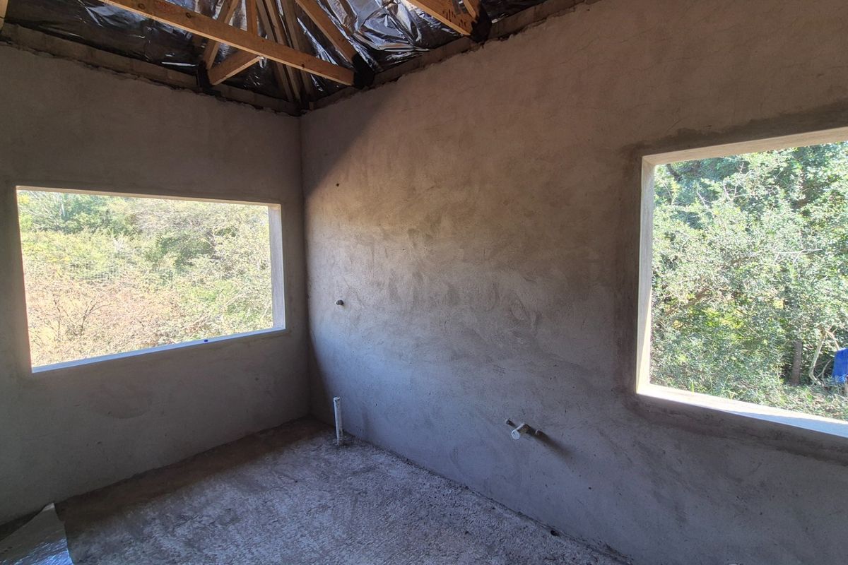 An unfinished bedroom space in the construction phase, featuring exposed roof trusses, plastered walls, and concrete flooring with plumbing rough-ins visible.