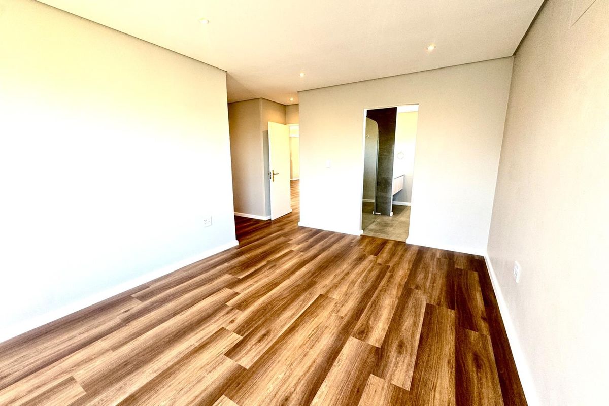 A bright, empty bedroom featuring modern laminate flooring, recessed lighting, and a doorway leading to an adjoining bathroom area.