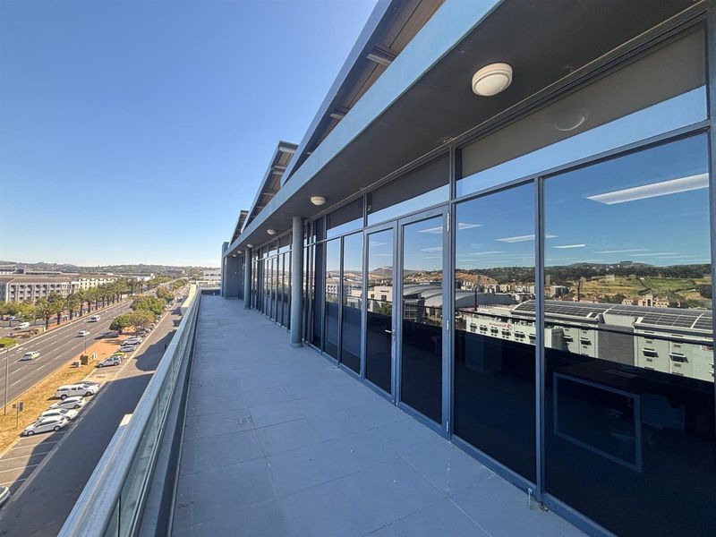 A spacious, modern office balcony featuring a glass balustrade, tiled flooring, and extensive floor-to-ceiling glass windows overlooking the surrounding urban landscape.