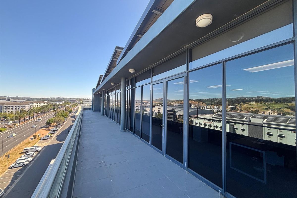 A spacious, modern office balcony featuring a glass balustrade, tiled flooring, and extensive floor-to-ceiling glass windows overlooking the surrounding urban landscape.