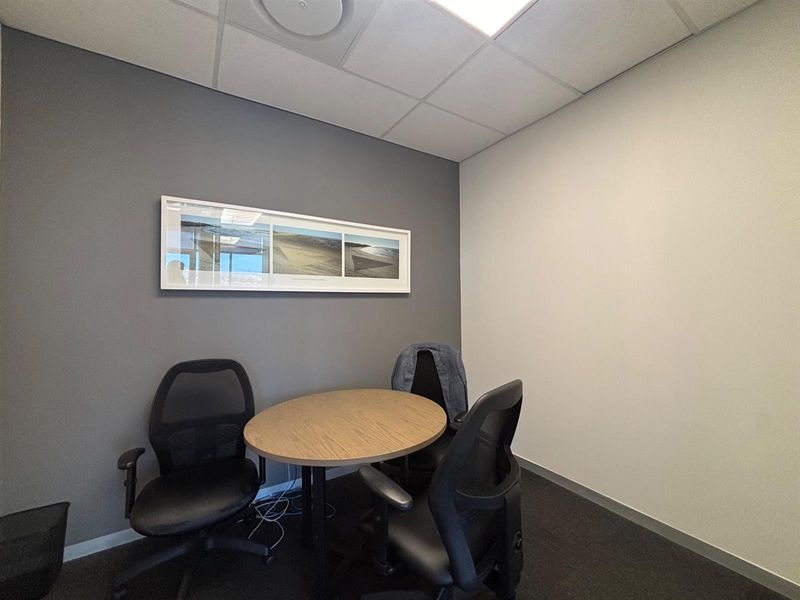 A compact, modern office space featuring neutral grey and white walls, carpeted flooring, and a suspended ceiling with recessed lighting.
