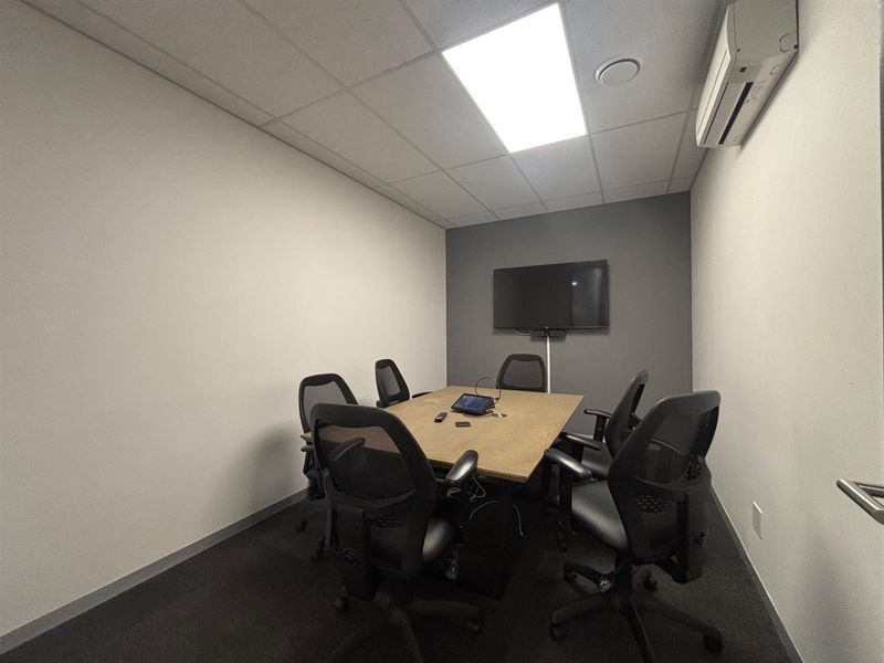 A functional office meeting room featuring a neutral color palette, carpeted flooring, recessed ceiling lighting, and a wall-mounted air conditioning unit.