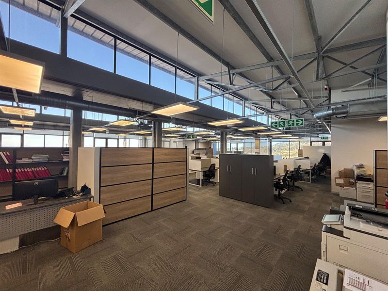 Spacious open-plan office area featuring high ceilings with exposed steel trusses, large clerestory windows for natural light, and durable carpet tile flooring.