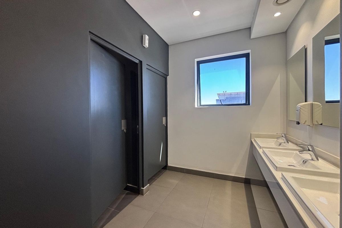 Modern office bathroom featuring a double vanity with ceramic sinks, chrome fixtures, large wall-mounted mirror, and neutral tiled flooring.