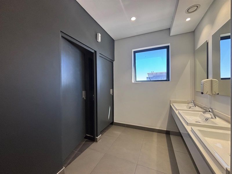 Modern office bathroom featuring a double vanity with ceramic sinks, chrome fixtures, large wall-mounted mirror, and neutral tiled flooring.