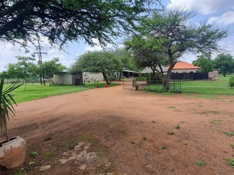 A wide view of the farm property entrance featuring a dirt driveway, established trees, and perimeter fencing leading toward the main dwelling.