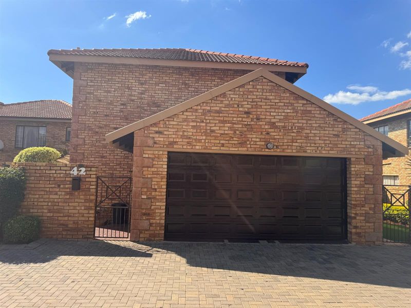 Front elevation of a face brick townhouse featuring a double garage, paved driveway, and secure pedestrian gate access.
