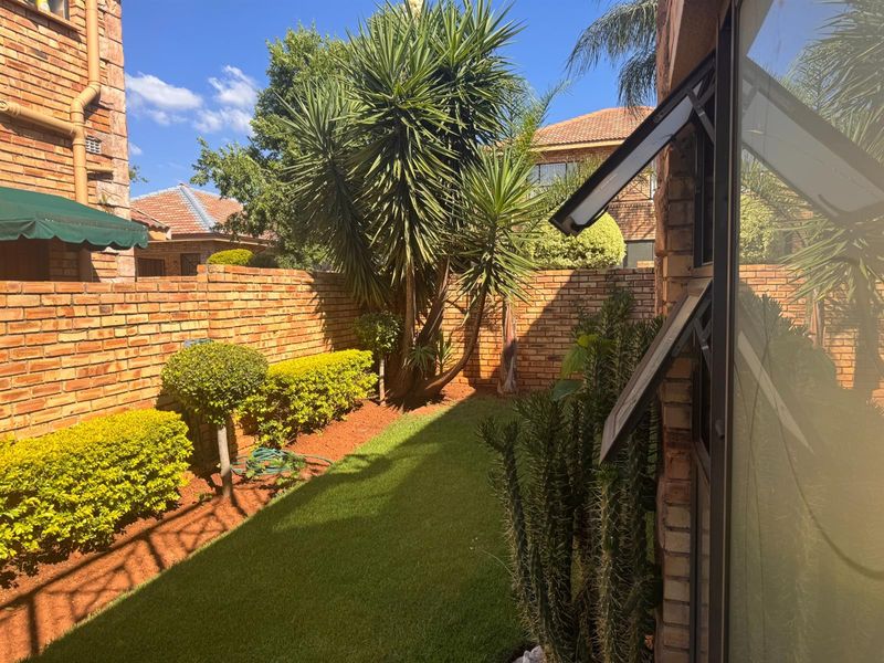 A private rear garden featuring a well-maintained lawn, established shrubs, and a face brick boundary wall, viewed from the side of the townhouse.