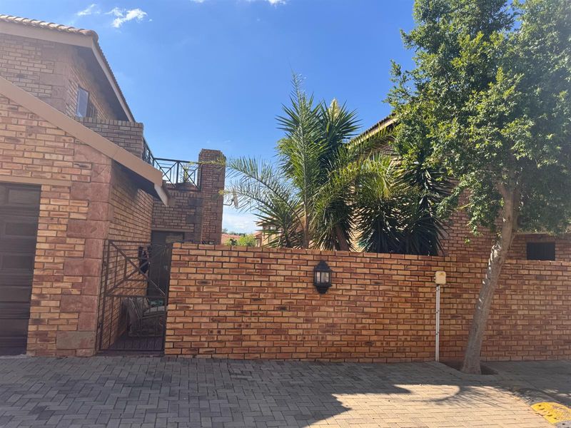 Front view of a face brick townhouse featuring a paved driveway, boundary wall, and an upper-level balcony.