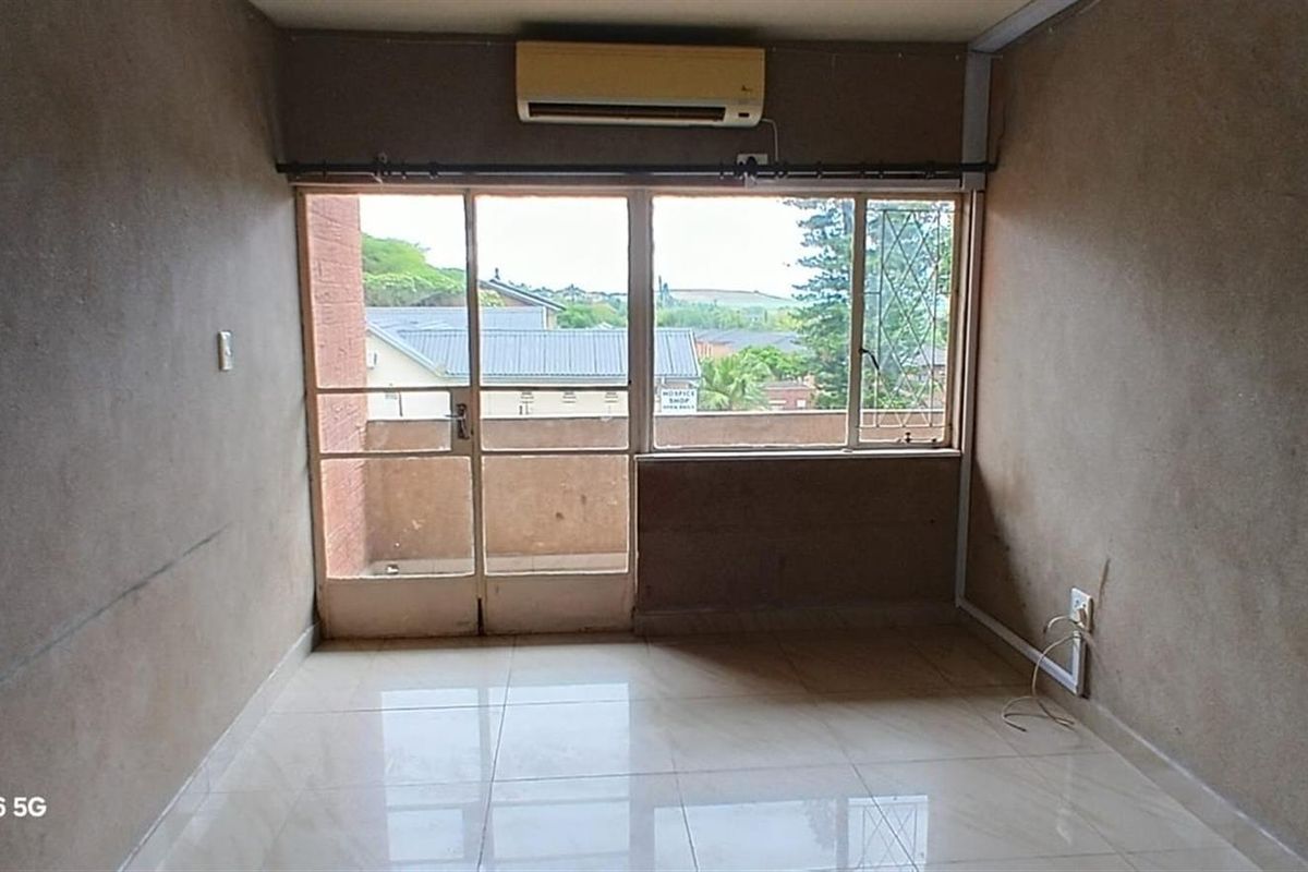 An empty bedroom featuring tiled flooring, a wall-mounted air conditioning unit, and large windows providing natural light and access to a small balcony.
