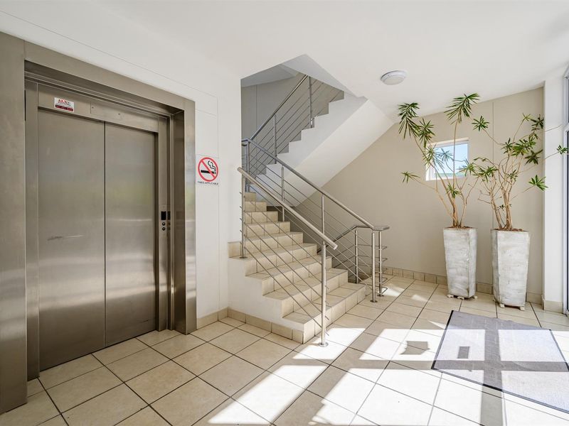 A clean and well-maintained apartment building lobby featuring a stainless steel elevator, tiled flooring, and a staircase with modern metal railings.