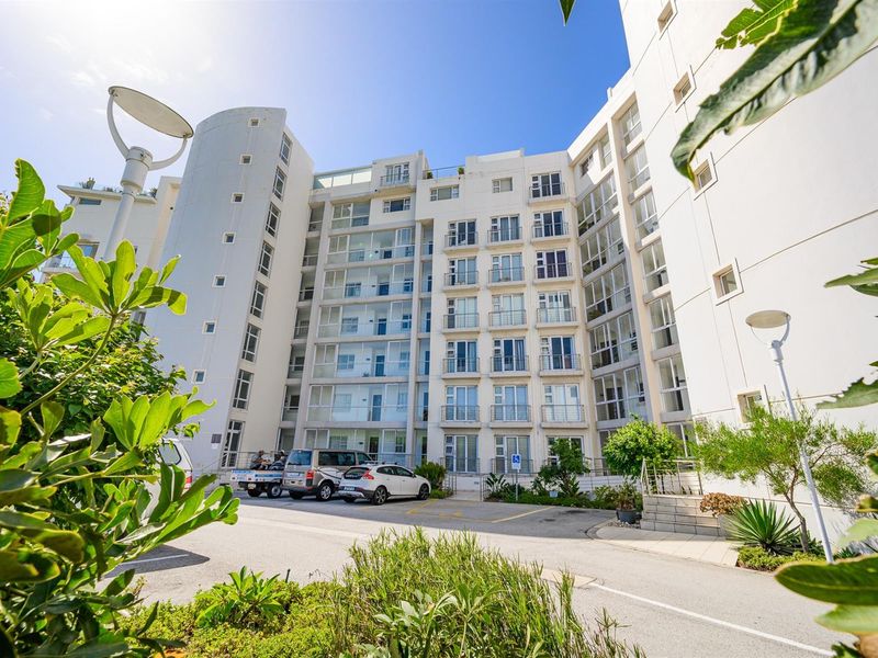 Exterior view of a modern multi-story apartment building featuring glass balconies and a paved entrance area with landscaped surroundings.