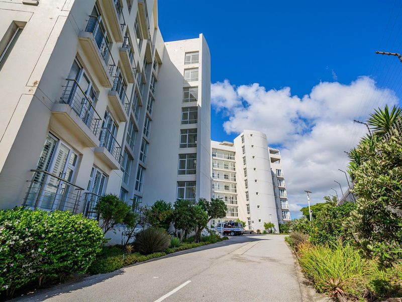 Exterior view of a modern multi-story apartment complex featuring white facade, glass balconies, and landscaped surroundings with paved driveway access.