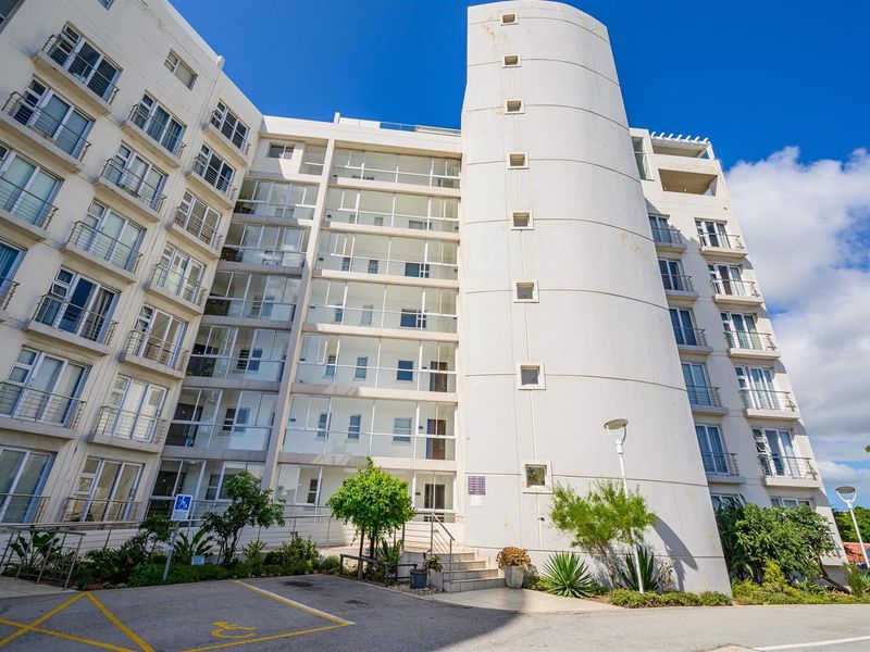 A view of the modern apartment building exterior, featuring a prominent curved architectural element, glass-enclosed balconies, and paved parking area.